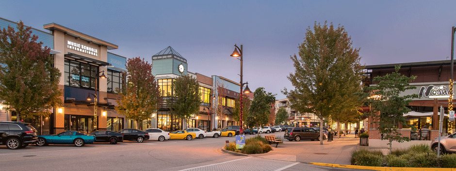Storefronts and street trees at Willoughby Town Centre in Langley at dusk, with parked cars along the street