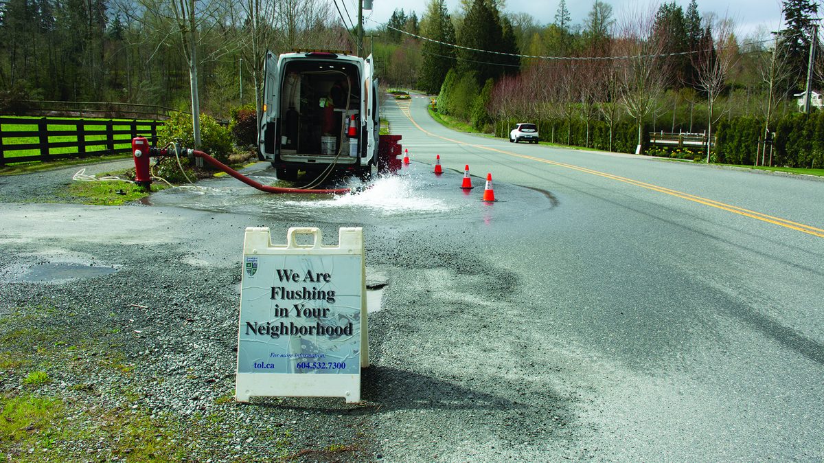 A Township crew van parked on a rural road with a red hose running from a fire hydrant, water gushing onto the pavement, traffic cones, and a sandwich board reading We Are Flushing in Your Neighborhood