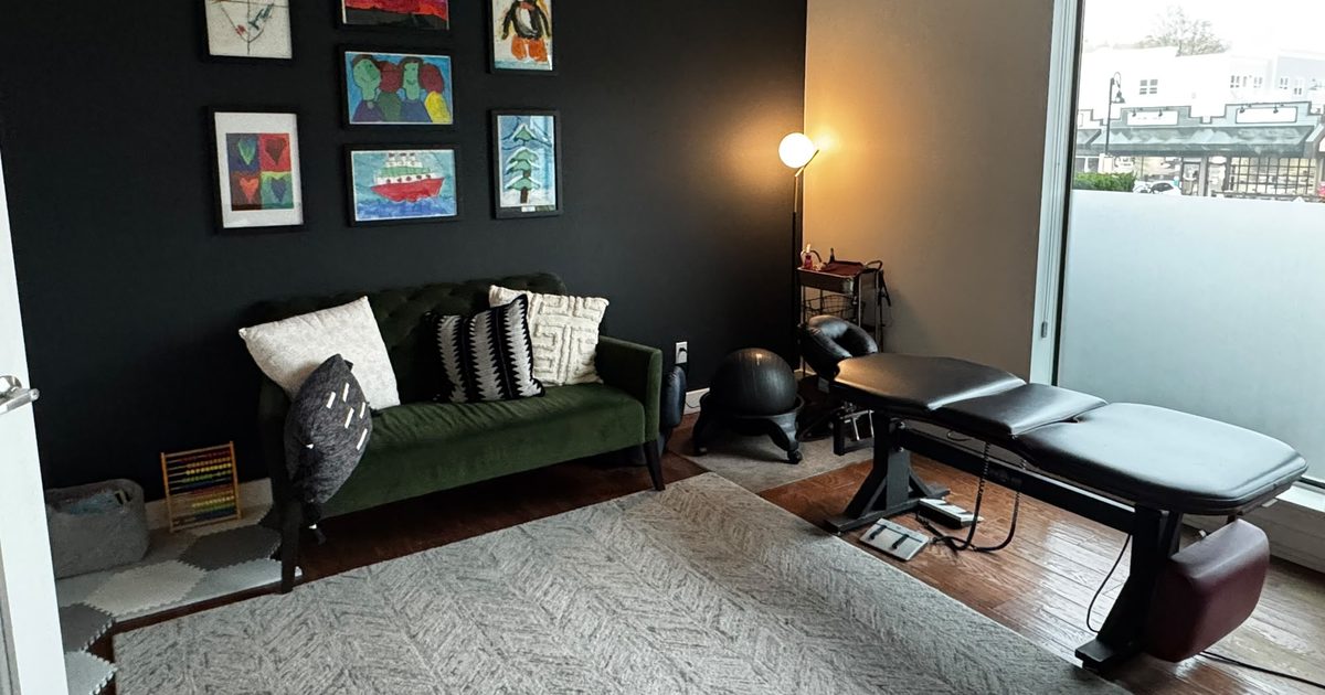 A Fort Family Chiropractic treatment room: framed kids' paintings on a black wall, a green velvet loveseat, a floor lamp and a black adjusting table by the window